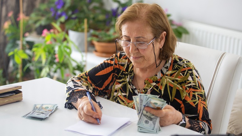 An older woman wearing glasses counts U.S. dollar bills while writing on a notepad at a table surrounded by houseplants