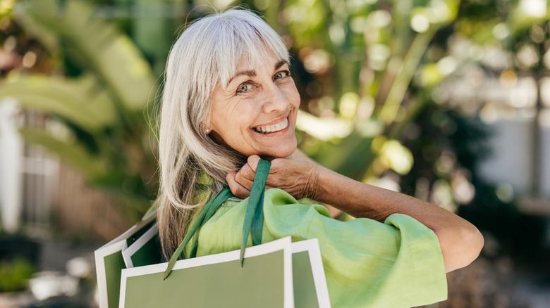 A mature woman with white hair smiles broadly with shopping bags draped over her shoulders, in front of a lightly blurred palm tree
