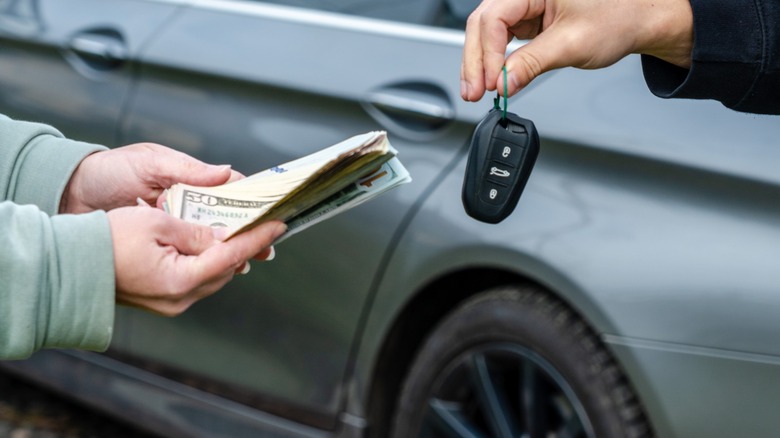 Close-up of a person handing over cash while another hands them a car key fob in front of a parked car, suggesting a private vehicle sale