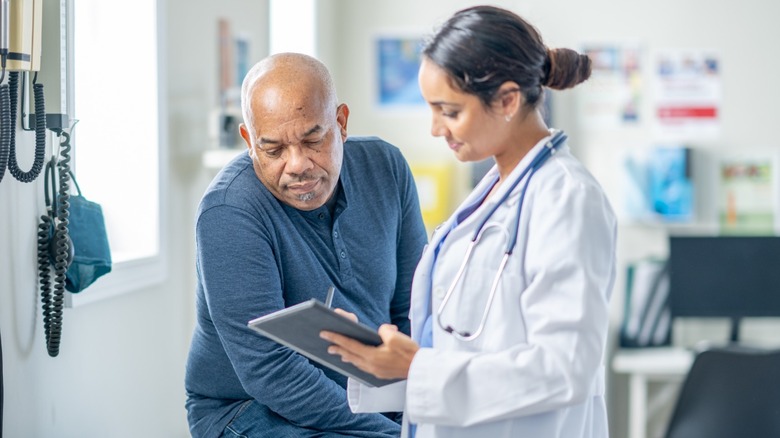 An older man sitting on an exam table as a doctor in a white coat reviews information on a tablet during a medical visit