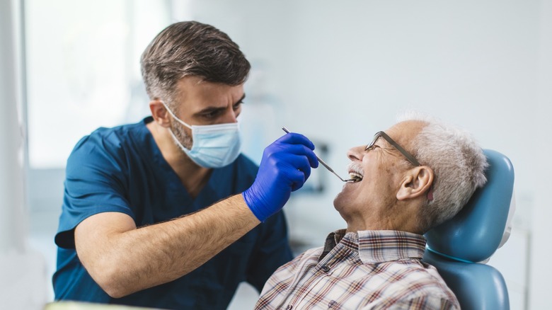 Dentist in scrubs and gloves examines an older man's teeth as he reclines in a dental chair during an appointment