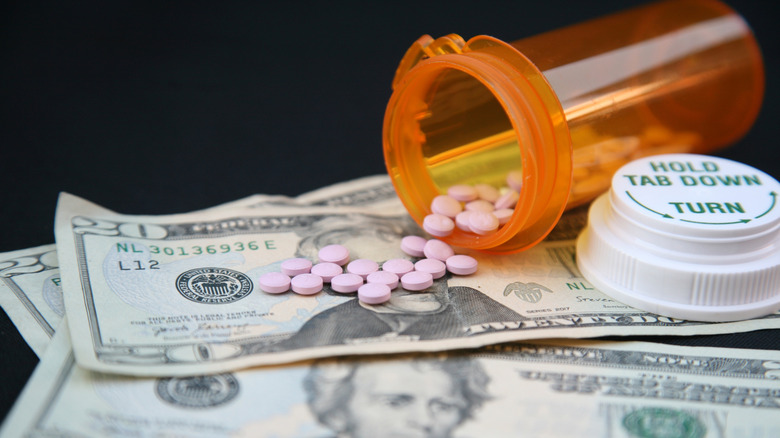 Opened pill bottle, with pills spilling on top of pile of money