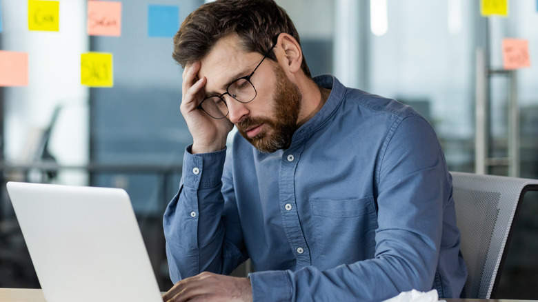 Stressed man wearing glasses sits at desk in front of his laptop