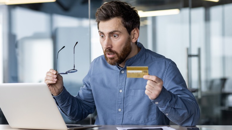 Shocked young male office worker sitting at workplace, holding wallet and credit card and looking worriedly at laptop screen.