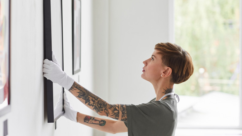 A woman with tattoos and wearing white gloves hanging a painting.