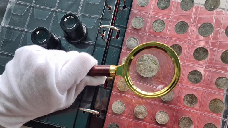 A gloved hand holding a magnifying lens over a book of old coins.