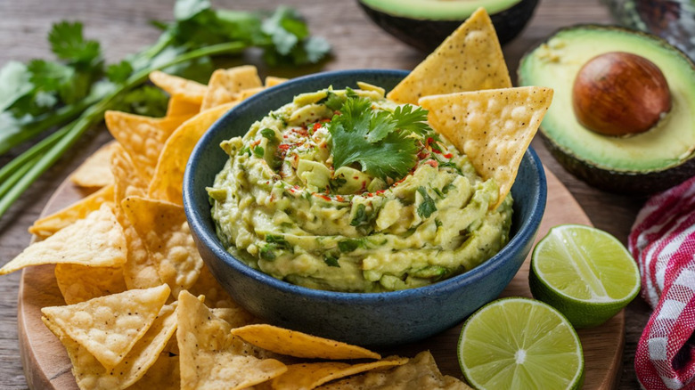 A bright green restaurant serving of guacamole in a bowl, surrounded by tortilla chips, limes, avocado, and herbs