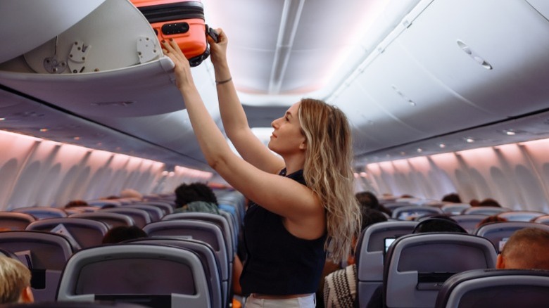 A female passenger putting a carry-on suitcase into the overhead compartment on a commercial plane.