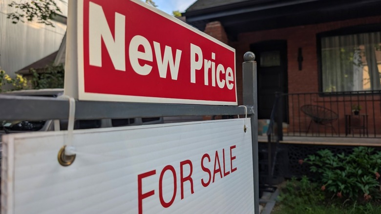 A new price sign on top of a for sale sign in front of a brick house with a porch that is having difficulty selling.