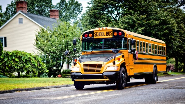 A typical yellow school bus driving down a residential street with a brick house visible.