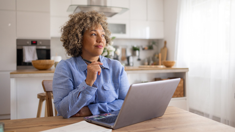 A middle-aged woman sitting at her kitchen table with a laptop looking thoughtful and holding a pen.