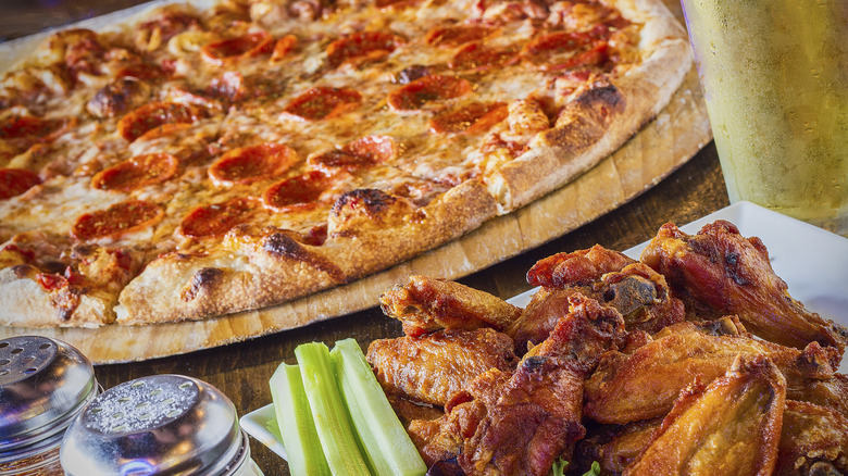 A pile of chicken wings next to a pepperoni pizza.