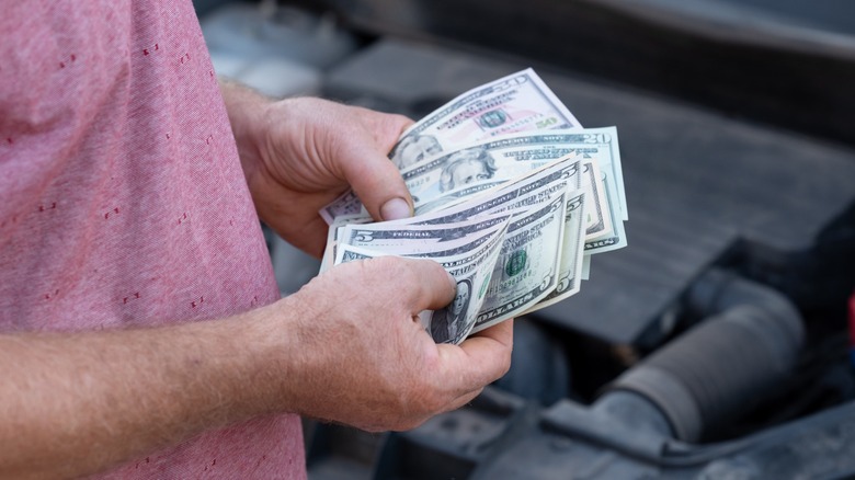 A person holds several U.S. dollar bills while standing beside an open car engine bay