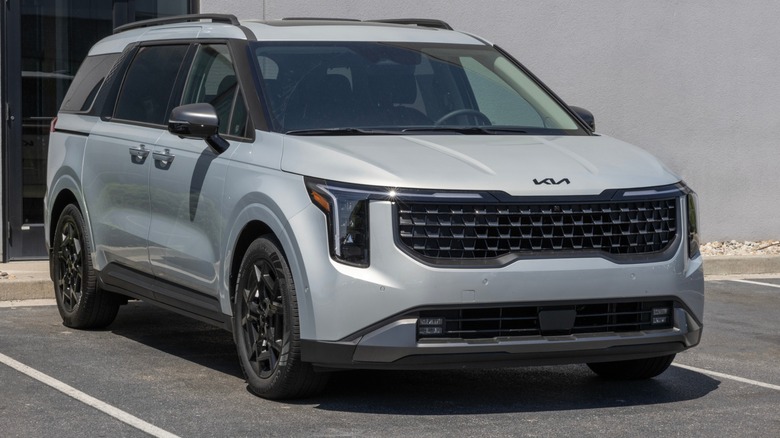 Silver Kia Carnival minivan parked in a lot, shown from the front three-quarter angle near a building