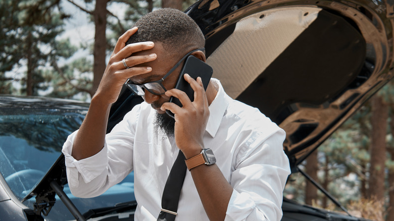 Man calling for assistance on his cellphone next to the open hood of an SUV