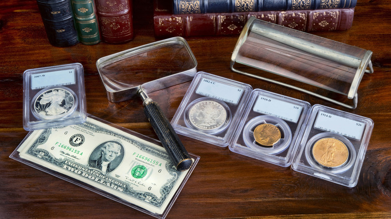 Coins and a two-dollar bill in protective cases on a table with a magnifying glass and books.