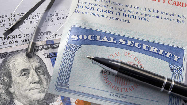 A Social Security card with a pen laying across it and money and paperwork in the background along with a pair of glasses barely visible in the corner.