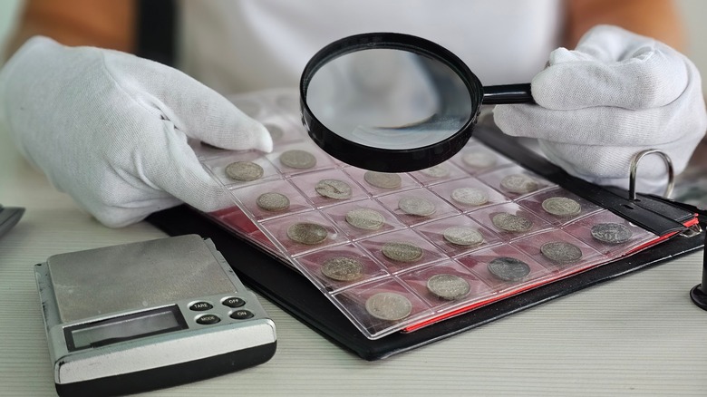 Picture of a faceless someone with a white shirt and gloves looking at a laminated book of coins. In one of the gloved hands is a magnifying glass.
