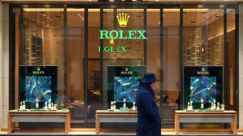Man in a trench coat stands outside a Rolex store