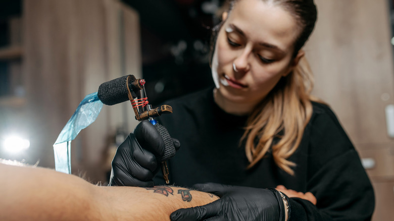 Young woman professional tattoo artist in black gloves making a rose tattoo on a man's forearm.