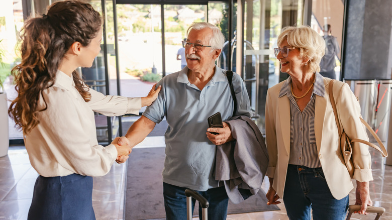 An elderly couple in casual travel attire stands in a hotel lobby