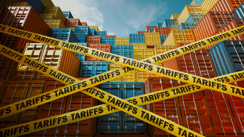 Yellow tapes with words "Tariffs" are stretched in front of a mountain of cargo containers.