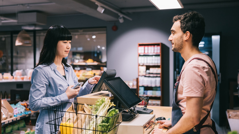 A young male cashier waits as a woman in business attire makes a payment at a grocery store
