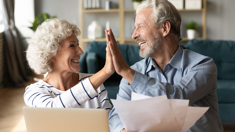 A happy retired couple giving each other a high five with the man holding papers and a laptop in front of them.