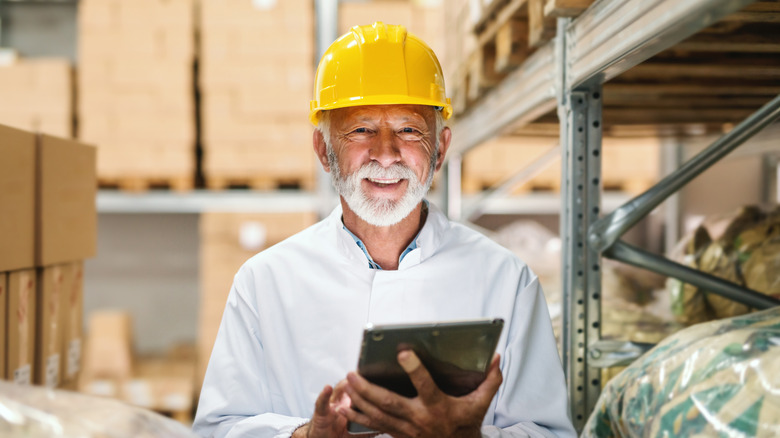 An older man wearing a hard hat in a warehouse holding a tablet.