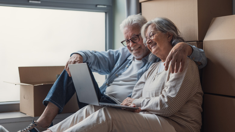 Older man and woman looking at open laptop while sitting on a floor in front of cardboard boxes