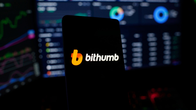 A Bitthumb logo on a black phone screen with trading metrics on a monitor in the background