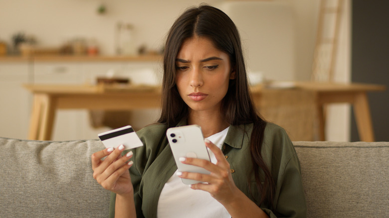 A woman with a serious look sitting on a couch holding a credit card in one hand a a cell phone in the other hand.