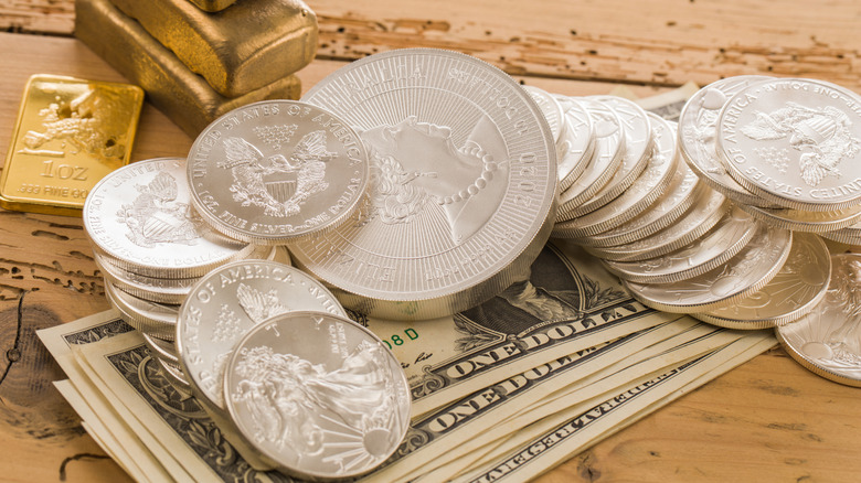 Silver coins piled on top of dollar bills with gold bars in the background on a wood table.