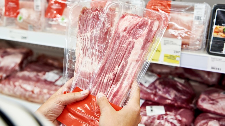 Hands holding two packs of sliced raw bacon in a grocery store meat aisle