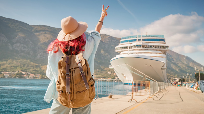 A woman traveler throwing up a peace sign in the direction of a docked cruise ship.