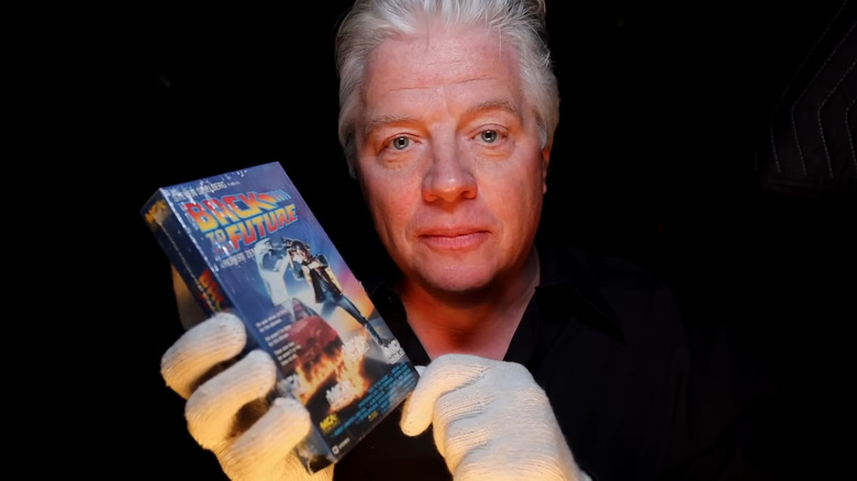 Actor Tom Wilson, wearing white gloves, holds up a sealed VHS of Back to the Future in front of a black background.