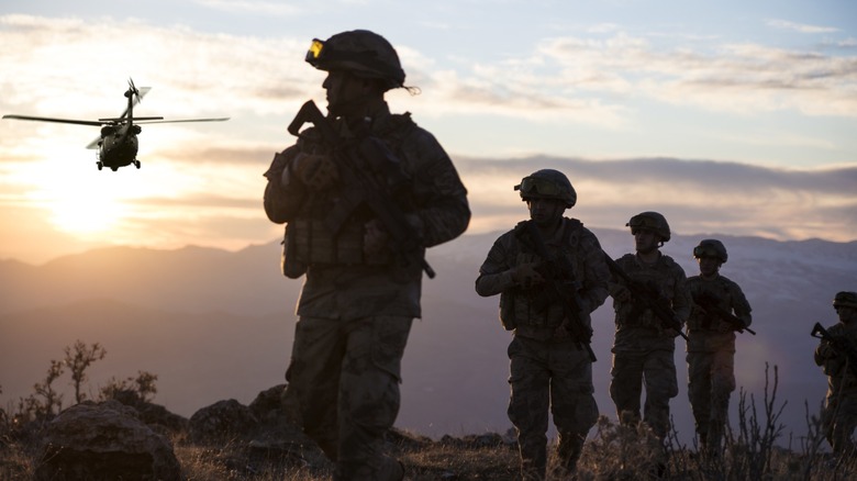 Members of the U.S. military armed and walking on a hill at sunrise with a helicopter behind them.