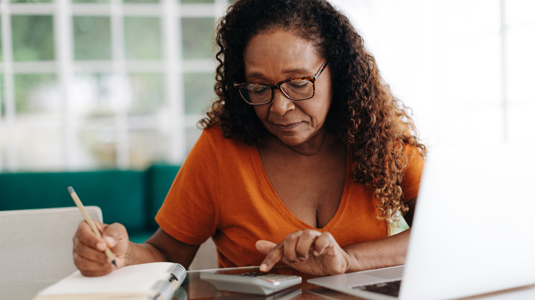 A senior woman reading a financial document at home in front of a laptop.