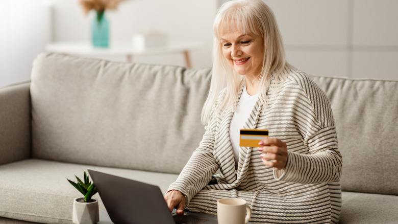 older woman with white hair smiling at laptop and holding credit card