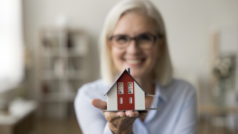 An blurred older woman holding out her hand with a miniature house on it.