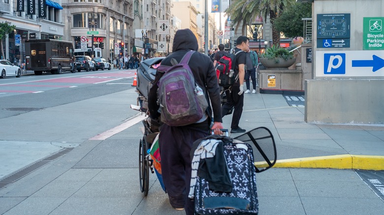 Man in hoodie wearing back pack carrying multiple pieces of luggage crosses street