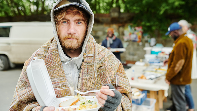 Disheveled man with beard holding container of food and plastic fork