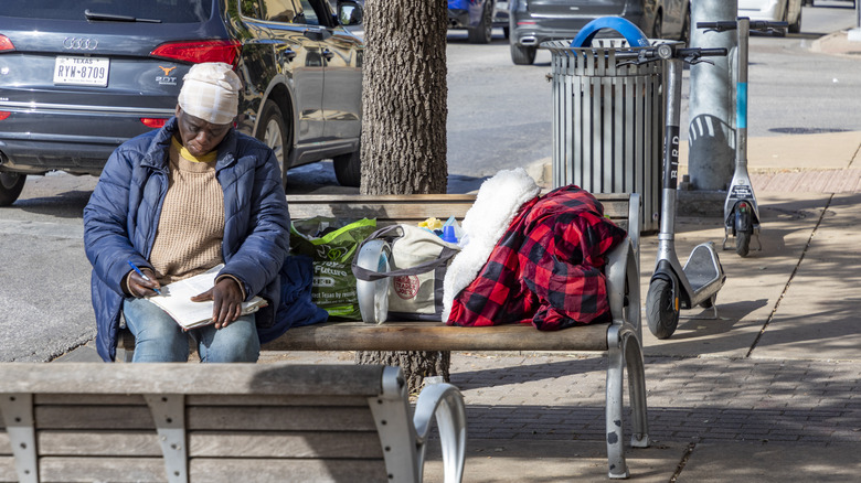Man sits with bags on city bench near street while cars drive by