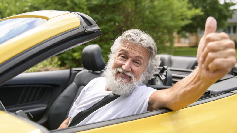 A smiling older man gives a thumbs-up while sitting in the driver's seat of a yellow convertible with the top down