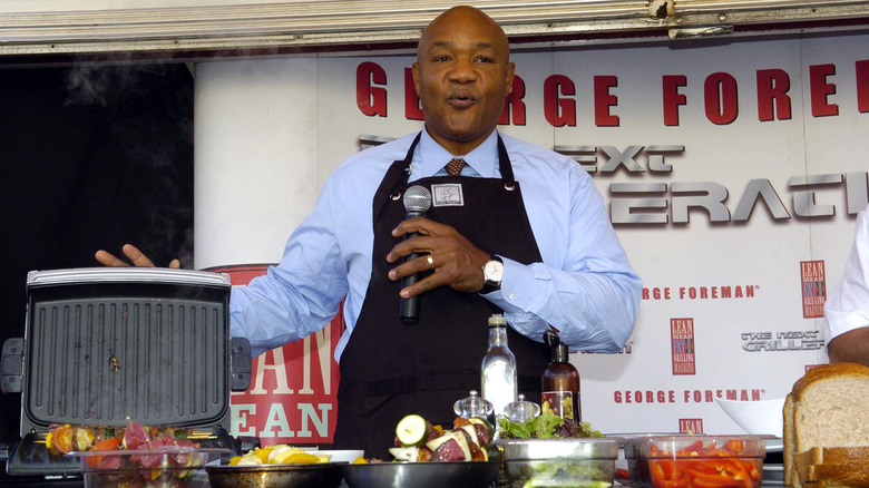 George Foreman standing next to the Foreman grill while speaking into a microphone.