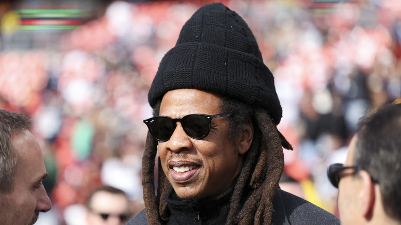 Jay-Z smiling while wearing shades in a black jacket and knit toque.