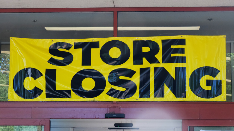 Store closing signs hanging inside of a defunct store with tall ceilings and fluorescent lighting.