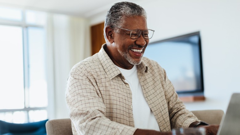An older man smiling while working on financial planning on a laptop in a living room.