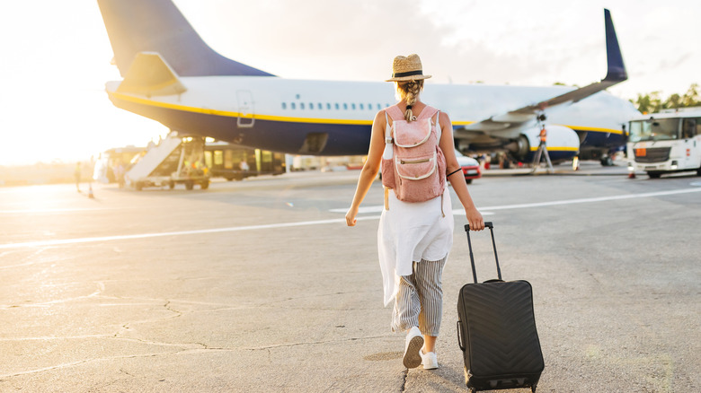 A female tourist walking toward a plane on the runway while dragging luggage behind her.