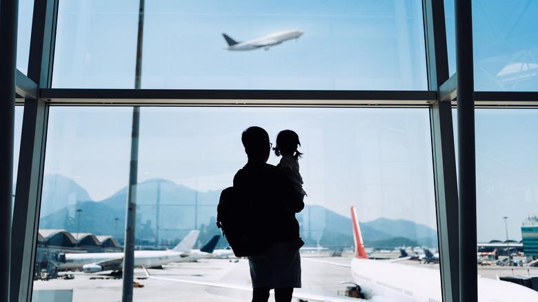 The silouette of a man carrying a little girl while watching a plane liftoff at the airport.